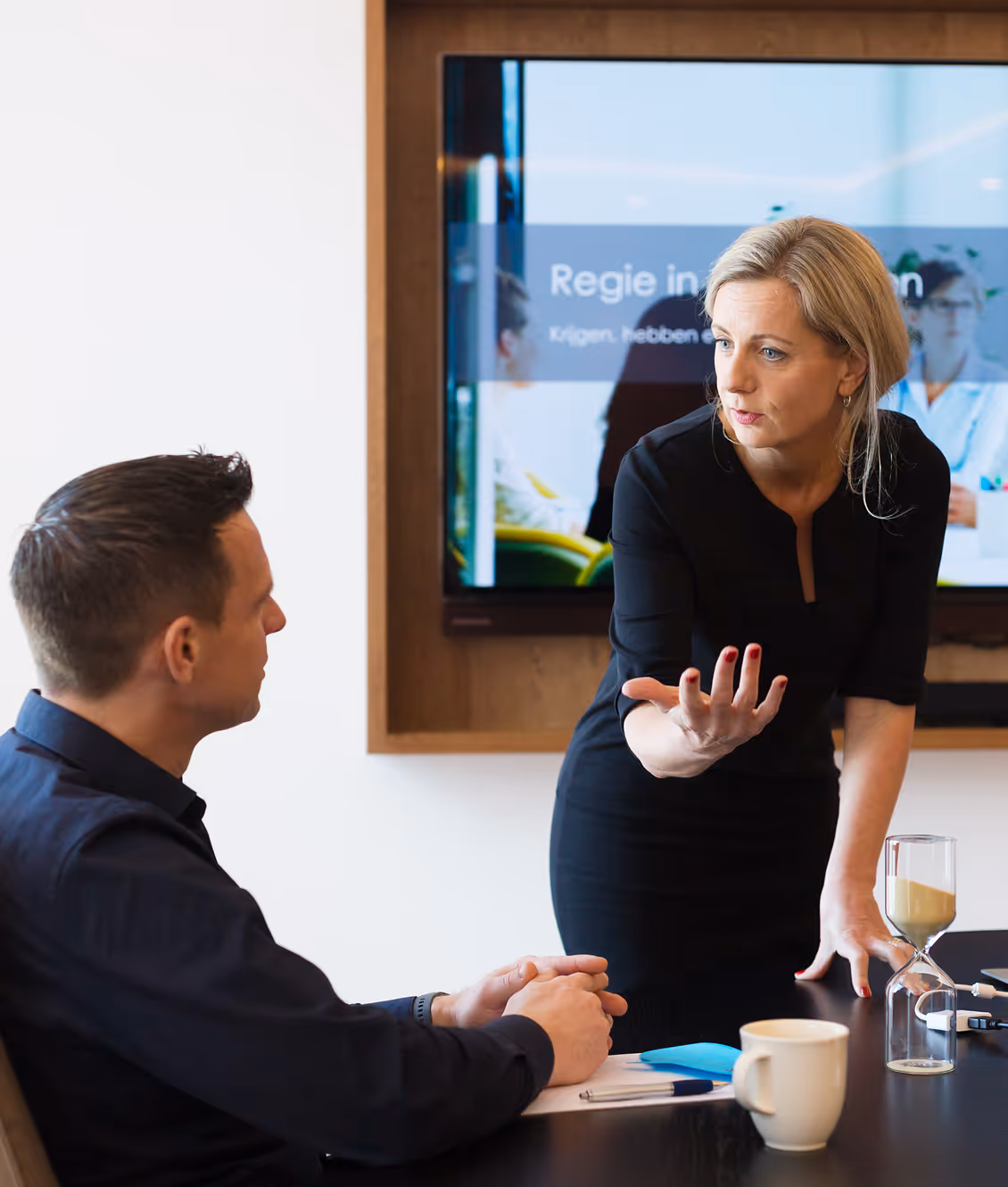 Woman in black dress talking and gesturing to a seated man during a meeting, with a screen in the background.