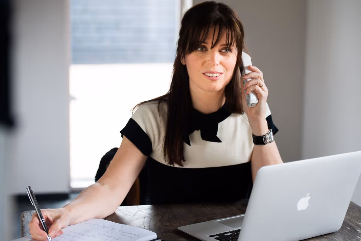 Woman with brown hair talking on a white phone while writing on paper at a desk with an open MacBook laptop.