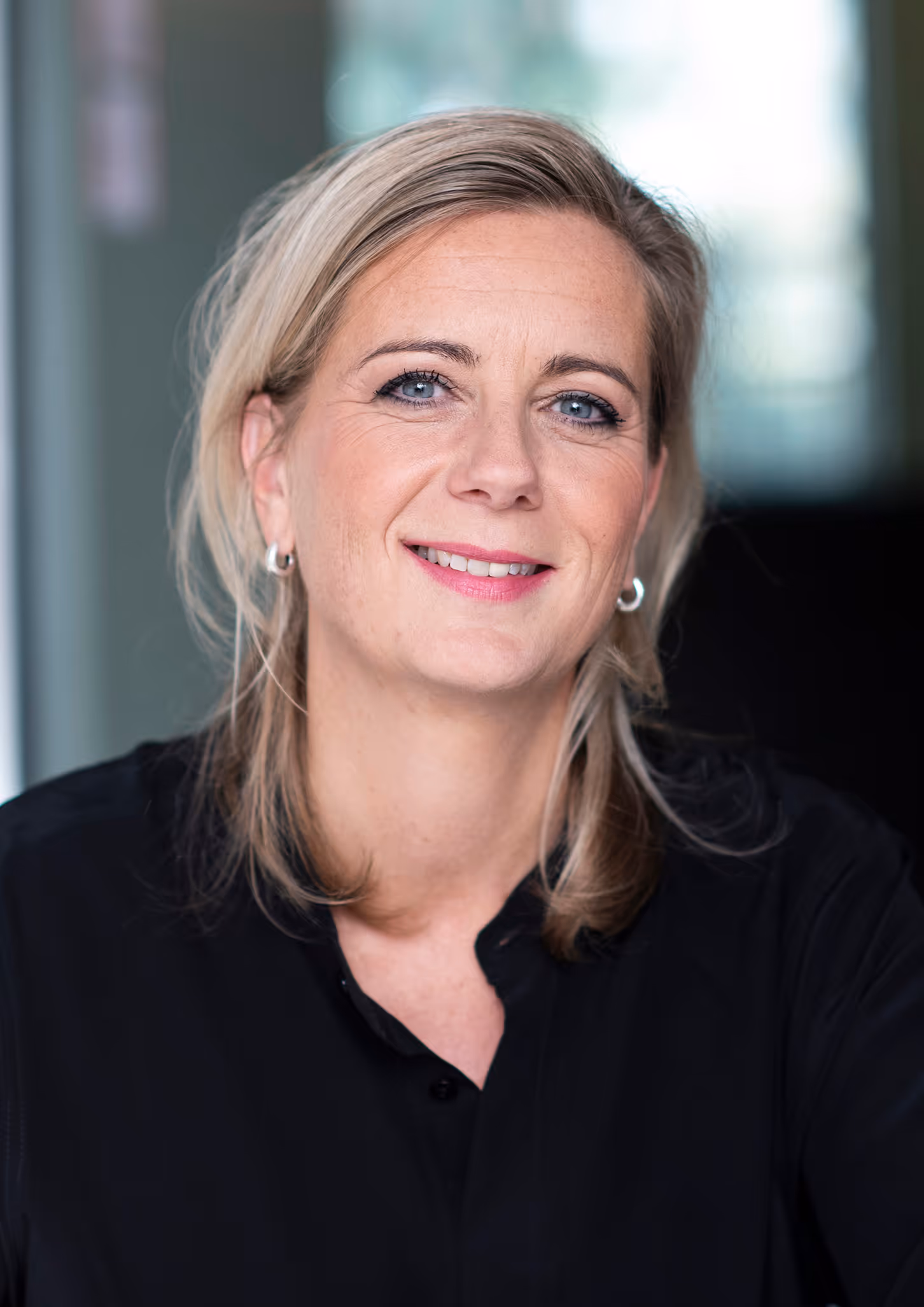 Smiling blonde woman with blue eyes wearing a black shirt and silver hoop earrings, against a blurred indoor background.