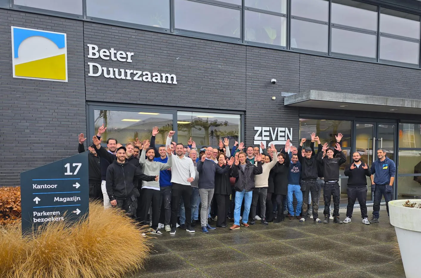 Group of diverse people smiling and waving in front of a building with the sign 'Beter Duurzaam' and a directional sign showing office, warehouse, and visitor parking.