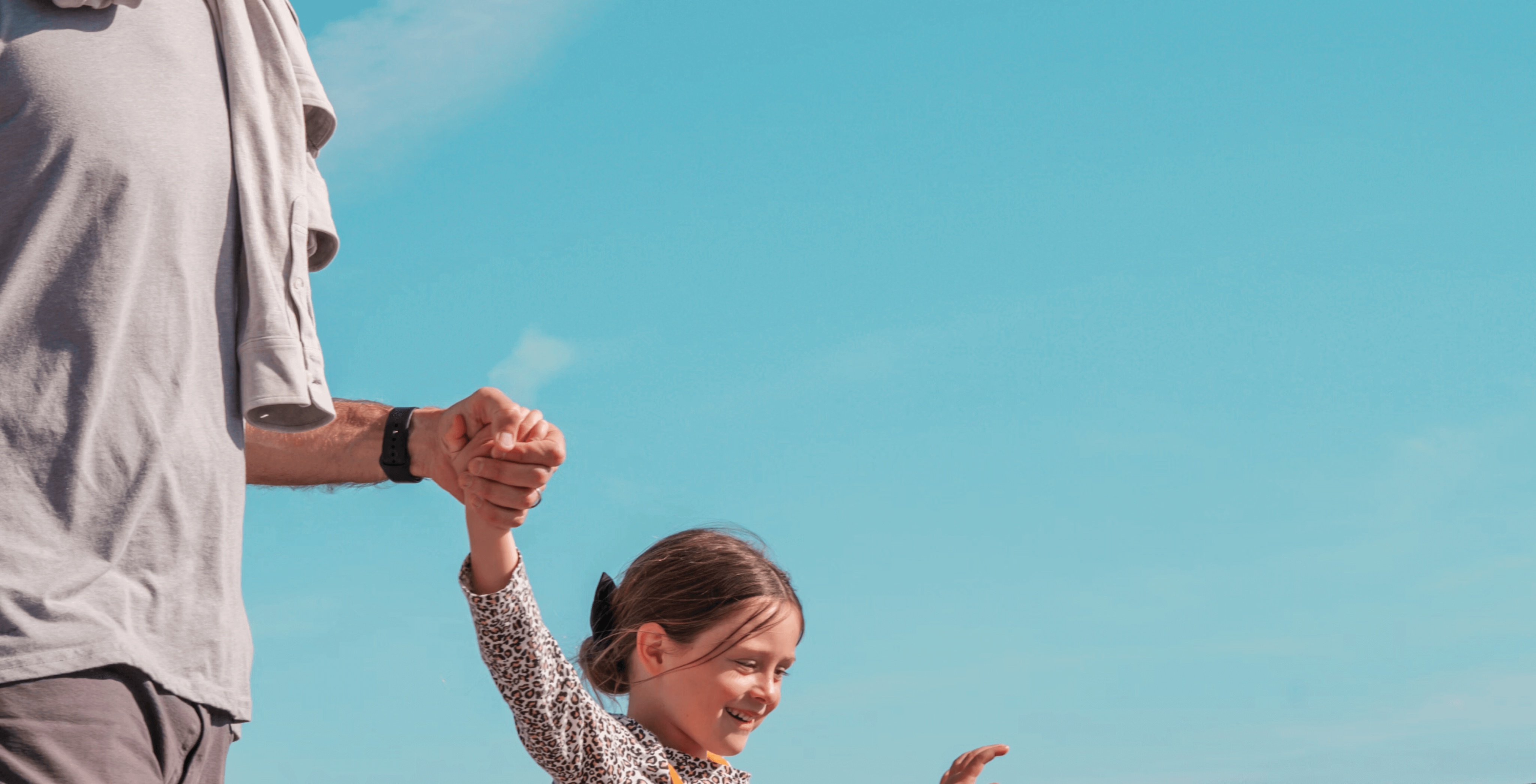 A man holding the hand of a smiling girl, who is joyfully reaching out her other hand. They are outside with a clear blue sky in the background, suggesting a playful moment together.