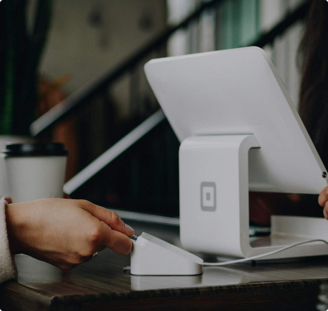 A close-up view of a payment terminal with a hand using a card reader beside a tablet. A coffee cup is visible in the background, indicating a cafe or small business setting. This scene suggests a transaction at Bonza Small Business Brokers.
