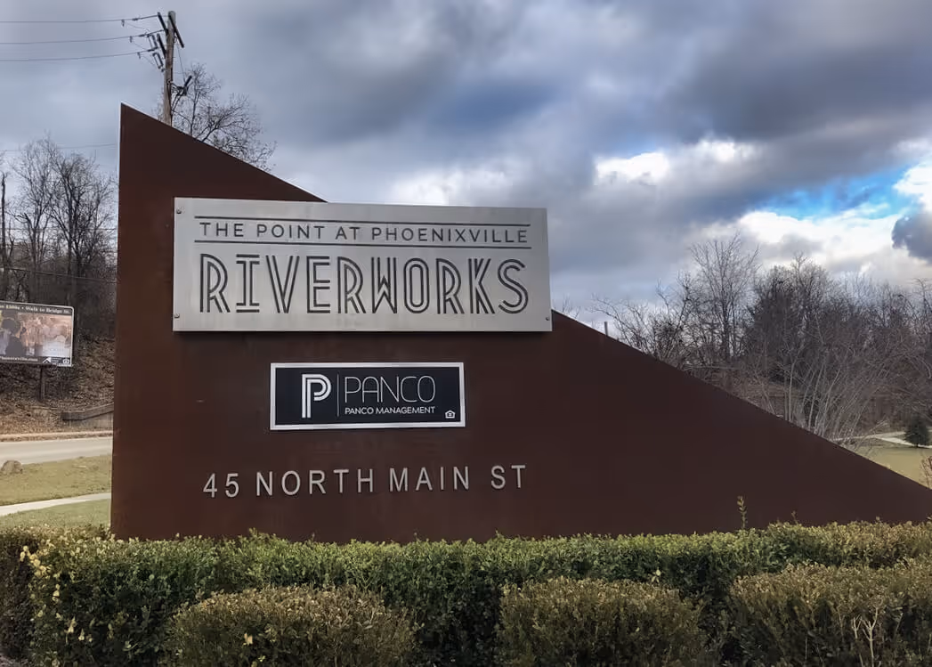 Tall triangular concrete monument with rectangular aluminum panel for location name whose top right corner is unattached to the concrete. Additional aluminum panel and text below for address and branding.