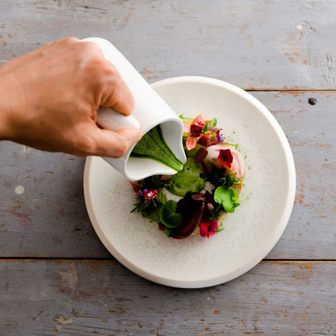 Ruka nalévá zelenou omáčku z bílé konvice na gurmánské jídlo zdobené jedlými květinami a zeleninou na bílém talíři.