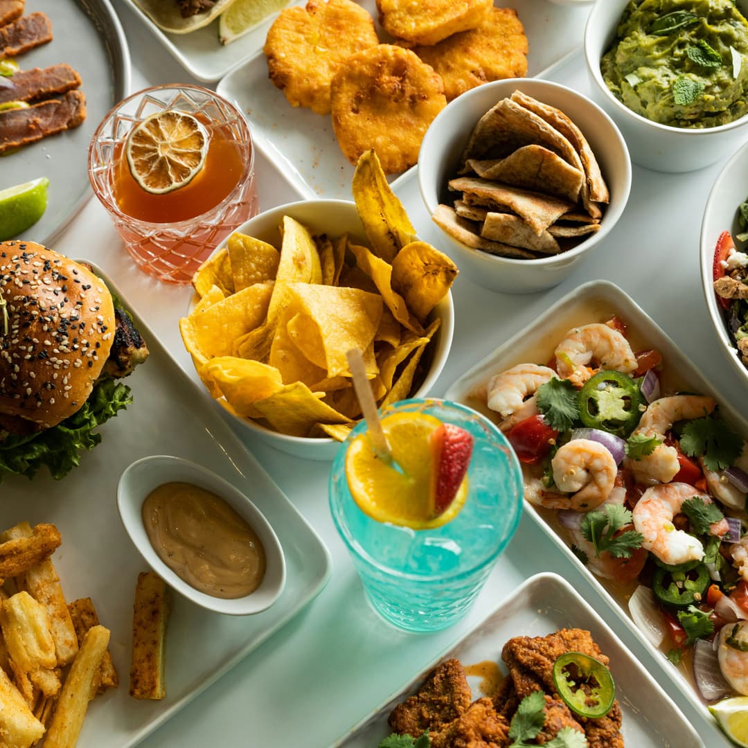 Různé pokrmy včetně krevetového ceviche s jalapeños, smaženého kuřete, chipsů s banánovými chipsy, guacamole, hamburgeru, smažených yucca hranolků a dvou barevných koktejlů.