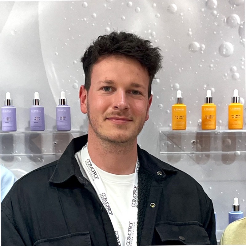 Muž s krátkými kudrnatými tmavými vlasy, oblečený v bílé košili a černé bundě, stojící před regály s fialovými a oranžovými lahvičkami s kosmetickými přípravky.