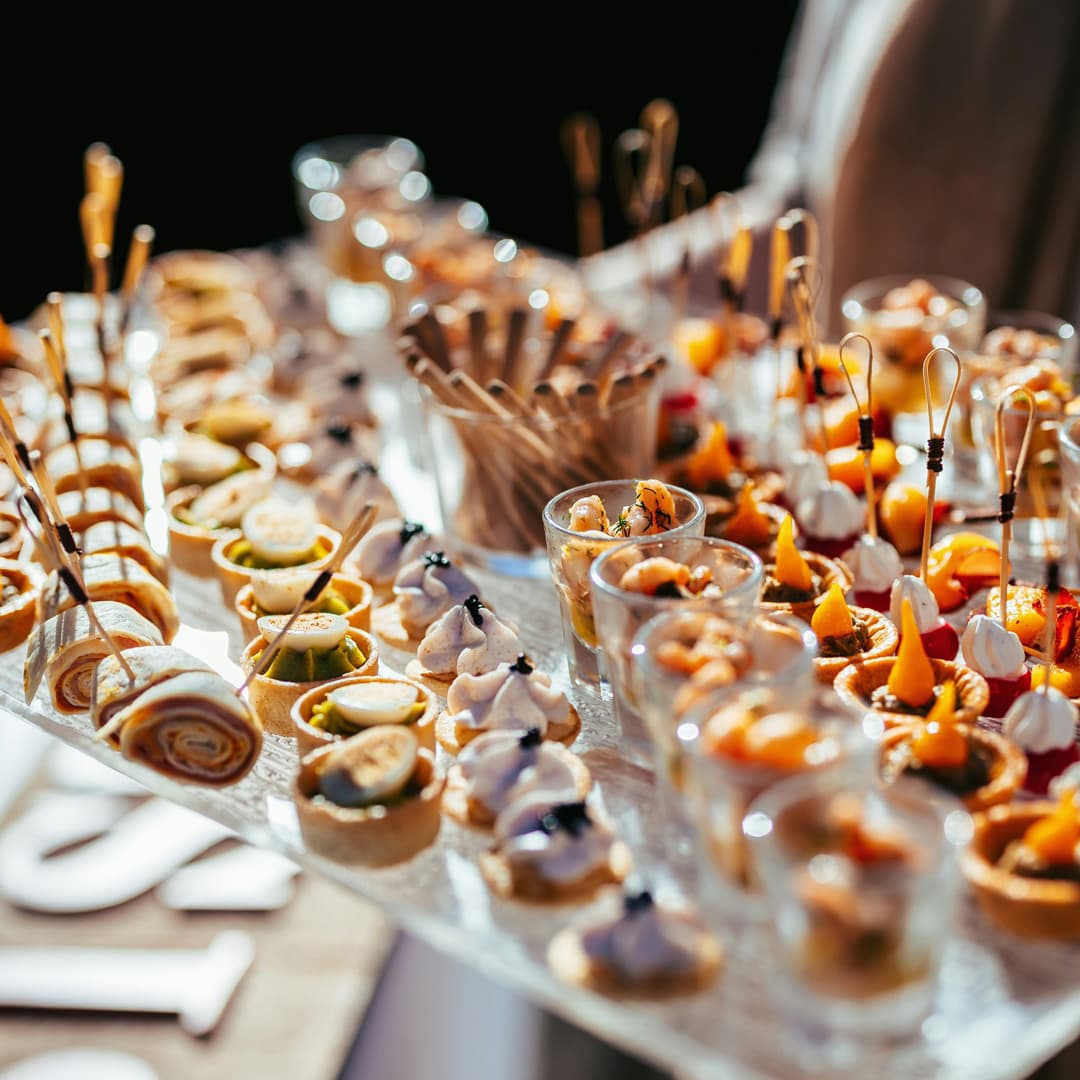 Různé elegantní jednohubky a předkrmy umělecky naaranžované na skleněném podnosu s špízy a malými skleničkami.