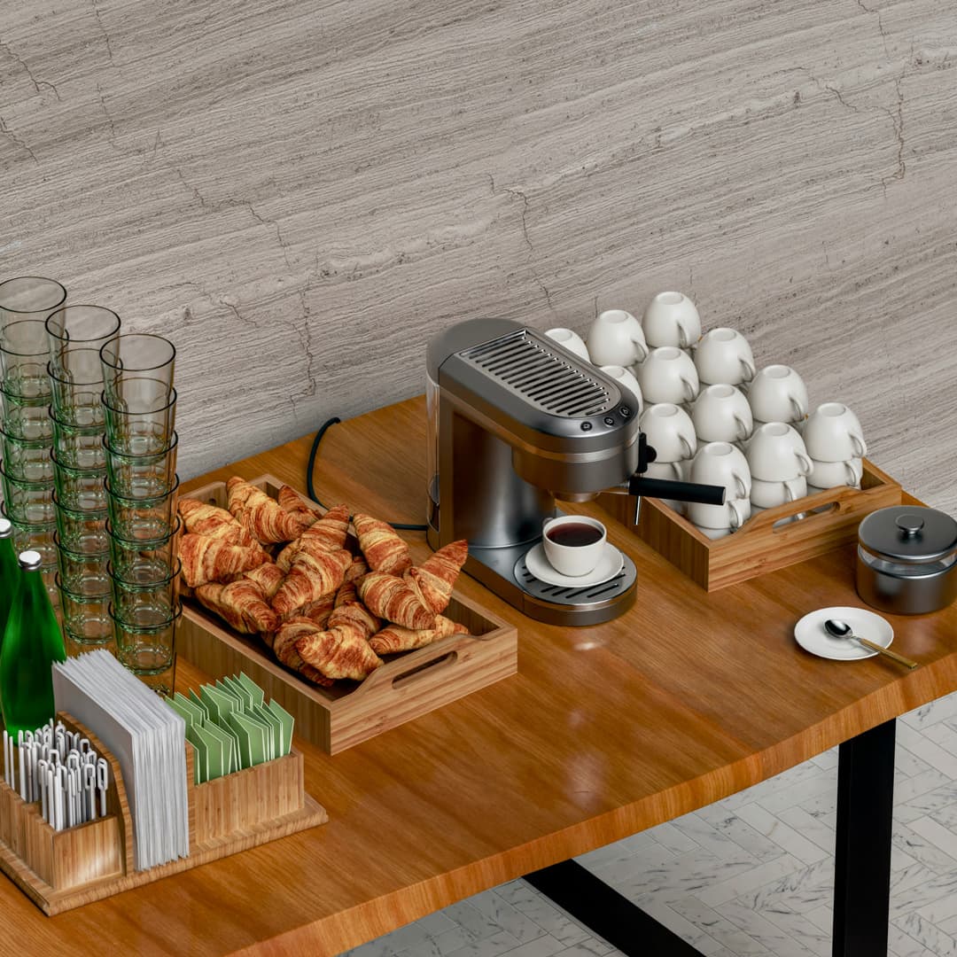 Stůl s kávovarem, bílými šálky, croissanty na dřevěných podnosech, naskládanými čirými sklenicemi, zelenou balenou vodou, ubrousky a míchátky před šedou texturovanou stěnou.
