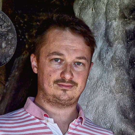 Muž s krátkými hnědými vlasy a vousy, oblečený v růžovo-bíle pruhovaném tričku s límečkem, stojící v interiéru u texturované šedé zdi.