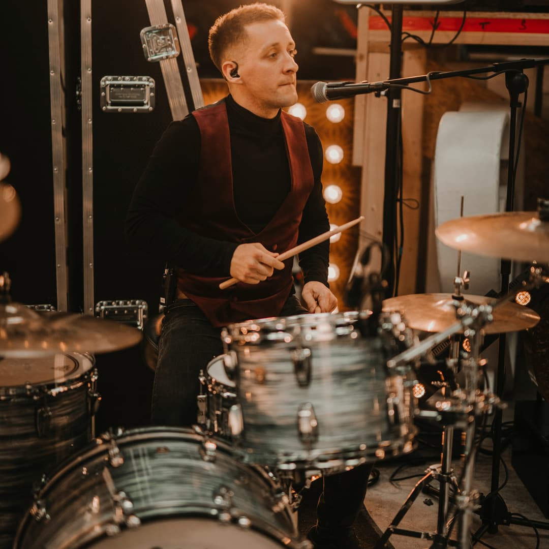 Bubeník v kaštanově hnědé vestě a černé košili hraje na bicí s mikrofonem před sebou.