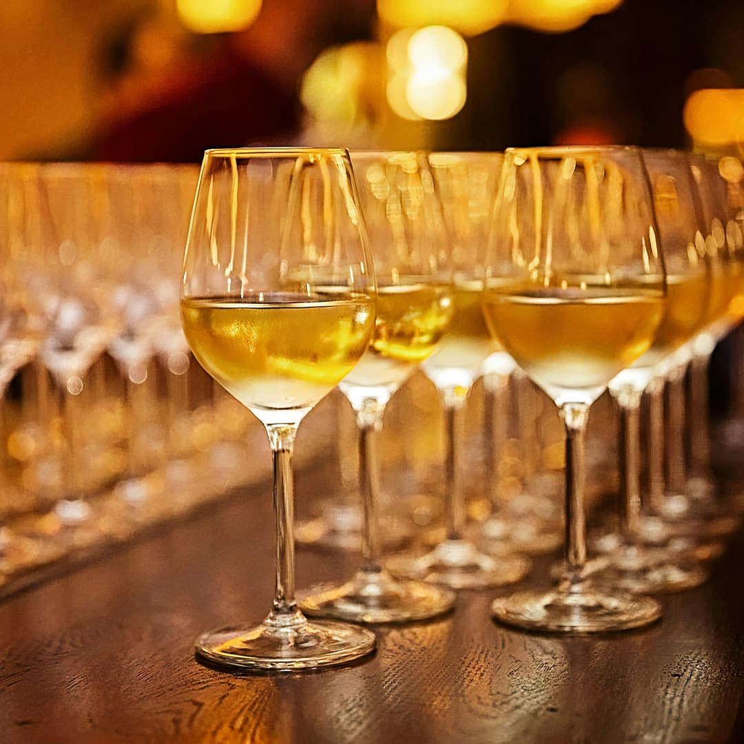 Řady sklenic naplněných bílým vínem na dřevěné desce pod teplým osvětlením.