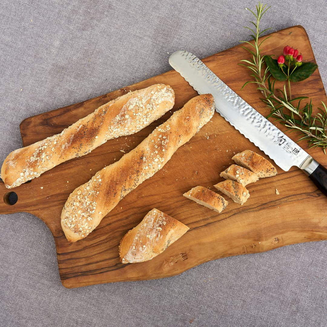 Dvě celé bagety s ovesnými vločkami a jedna částečně nakrájená bageta na dřevěné prkénko s vroubkovaným nožem, větvičkami rozmarýnu a malými červenými pupeny.