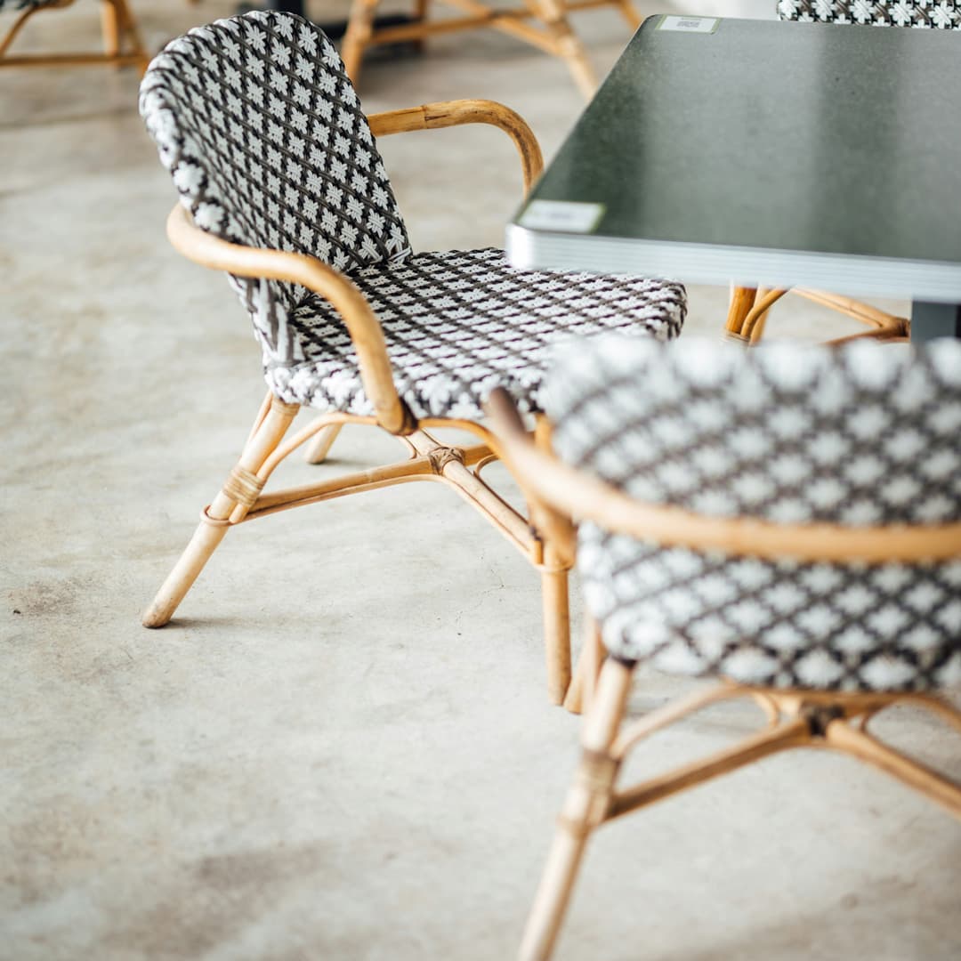 Detailní záběr proutěných židlí s černobílými vzorovanými polštáři vedle tmavého stolu na betonové podlaze.