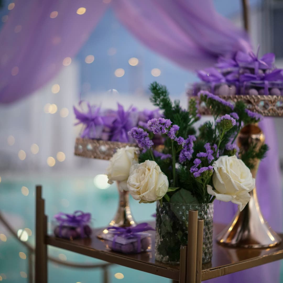 Bílé růže a fialové květiny v texturované skleněné váze na zlatém stole s fialově zabalenými dárky a jemně blikajícími světly v pozadí.