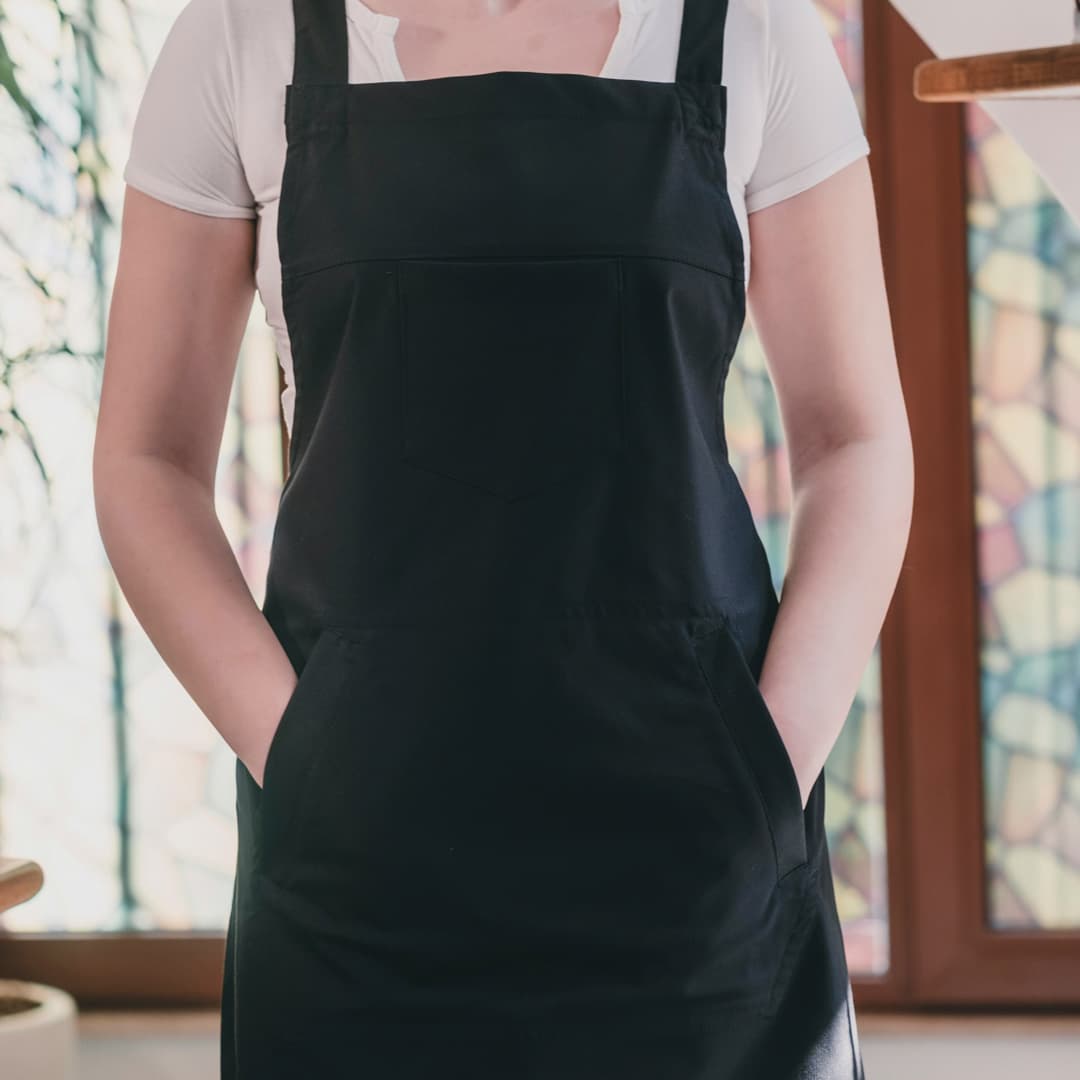 Osoba v černé zástěře přes bílou košili s krátkými rukávy, s rukama v kapsách zástěry, stojící v interiéru u okna s barevnými vitrážemi.