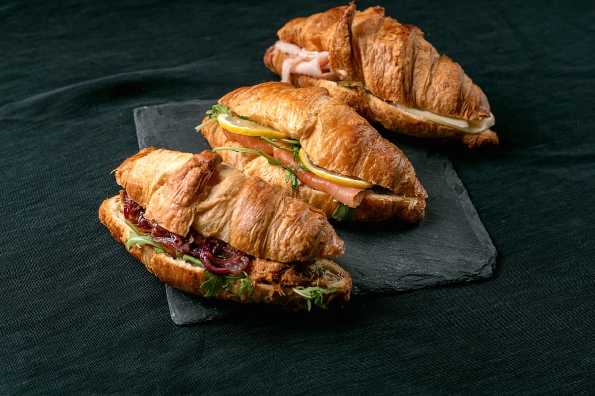 Tři croissantové sendviče s různými náplněmi na tmavé břidlicové desce a černém látkovém pozadí.