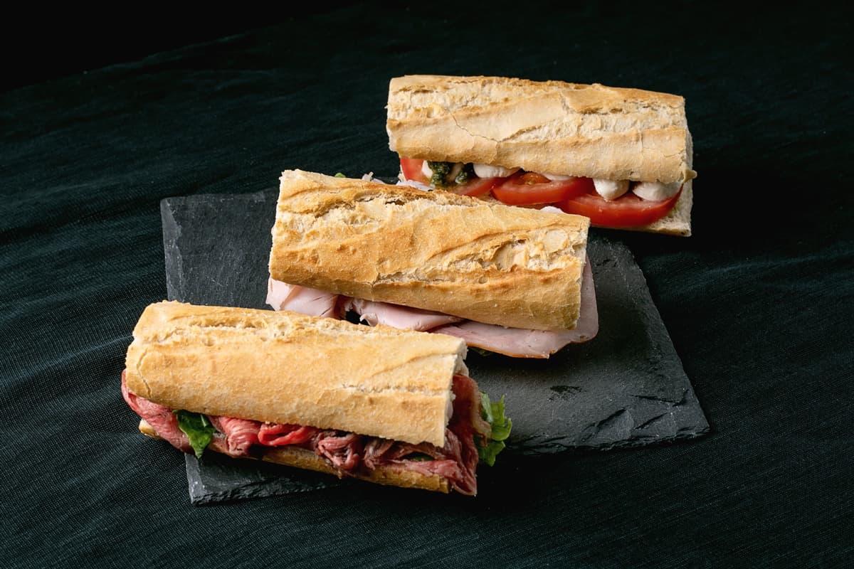 Tři bagetové sendviče s různými náplněmi, včetně pečeného hovězího masa, krůtího masa a rajčat s mozzarellou, podávané na tmavé břidlicové desce.
