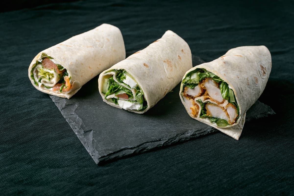 Tři tortillové wrapy s náplní včetně zeleniny, sýra, uzeného lososa a obalovaného kuřete, podávané na tmavé břidlicové desce.