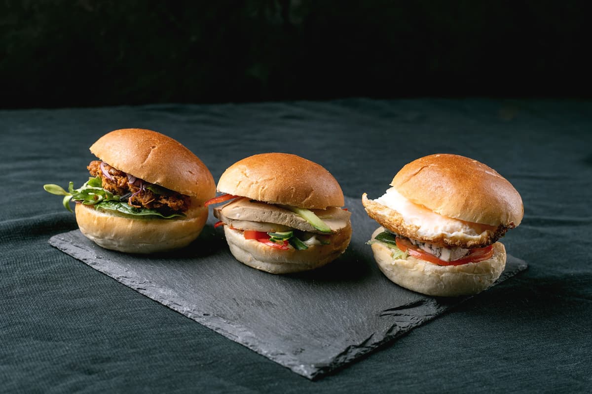 Tři mini sendviče s brioškovými houskami na podnosu z břidlice, smažené kuře, krůtí maso s avokádem a sendvič se smaženým vejcem a rajčetem.