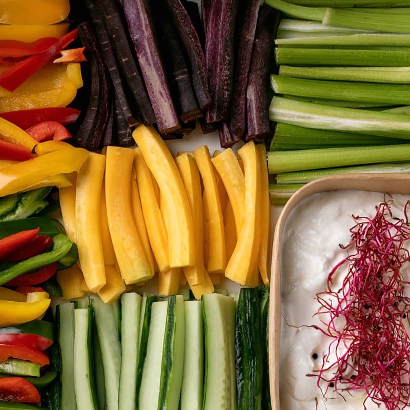 Různé čerstvé zeleninové tyčinky, včetně červené a žluté papriky, fialové mrkve, žluté cukety, celeru a okurek, naservírované kolem misky s bílým dipem a ozdobené červenými klíčky.