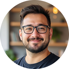 Usměvavý muž s brýlemi a vousy v rozmazaném interiéru.