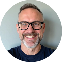 Usměvavý muž středního věku s brýlemi, krátkými šedivými vlasy a vousy, oblečený v tmavě modré košili, na světle šedém pozadí.