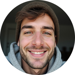 Mladý muž s vousy a knírem, na hlavě hnědou pletenou čepici a šedou mikinu s kapucí, usmívá se do kamery na tmavém pozadí.