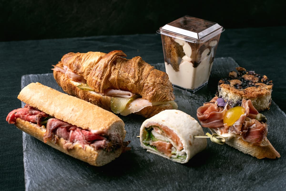 Různé gurmánské sendviče a dezerty na tmavém břidlicovém podnosu, včetně bagety s pečeným hovězím masem, croissantového sendviče, wrapu s uzeným lososem, bagety s prosciuttem a cherry rajčátky, parfaits v plastovém kelímku a borůvkového crumble baru.