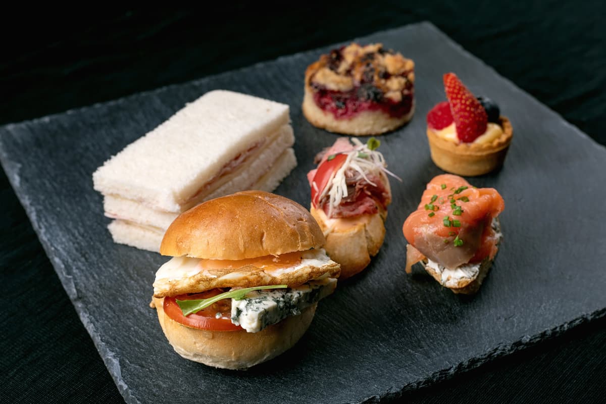 Různé malé sendviče a dezerty na černém podnosu, včetně sendviče s modrým sýrem, rajčaty, zeleninou a smaženým vejcem, mini sendviče, bruschetta s nakrájeným masem, uzený losos na toastu se smetanovým sýrem, bobulový koláč a ovocný koláč s jahodami a borůvkami.