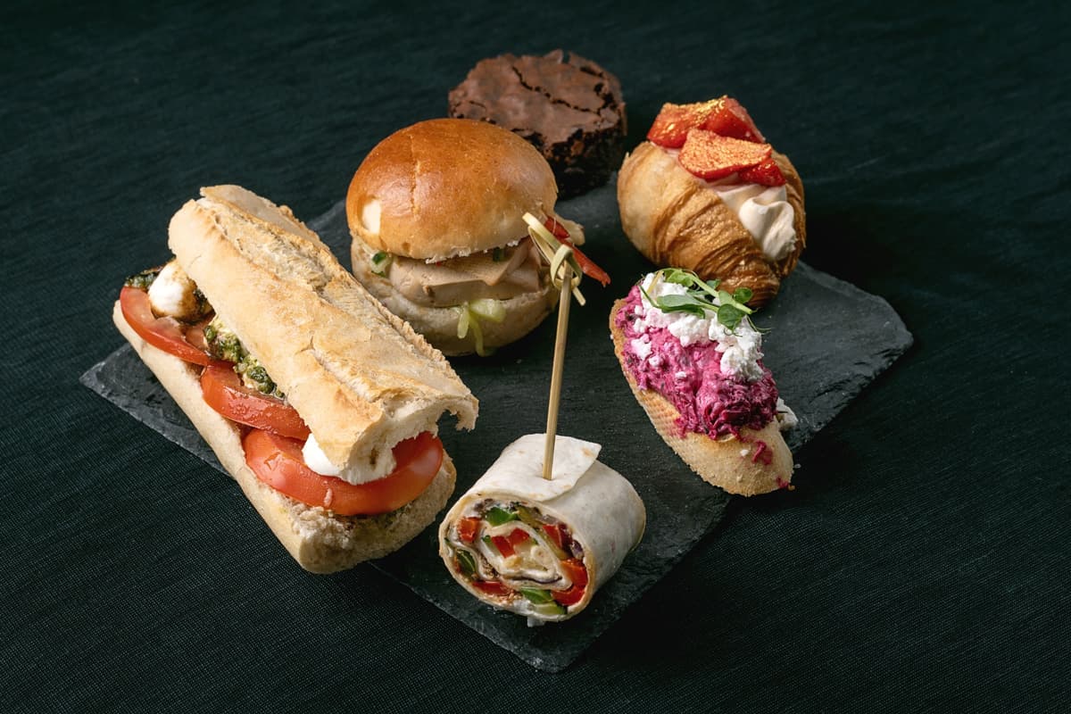 Různé gurmánské jednohubky, včetně bagetového sendviče s rajčaty, mini hamburgeru, wrapu, crostini s řepnou pomazánkou, croissantu se smetanou a jahodami a brownies na černém kamenném podnosu.