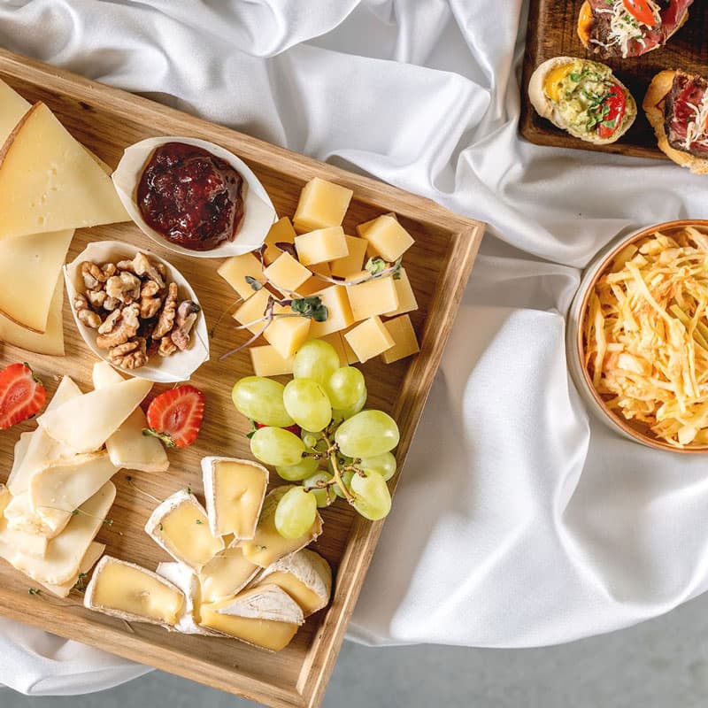 Dřevěný podnos s různými druhy sýrů, zelenými hrozny, vlašskými ořechy, marmeládou a nakrájenými jahodami na bílém ubrusu, vedle misky s nastrouhaným sýrem a malými bruschettami.
