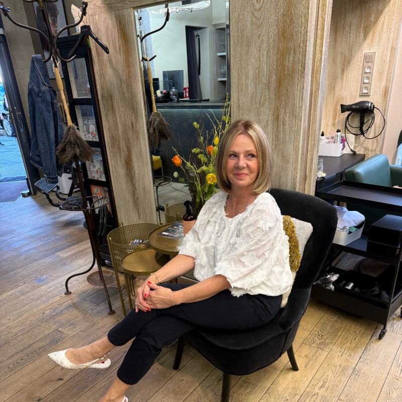 Žena středního věku s blond účesem, oblečená v bílé strukturované blůze a černých kalhotách, sedí na černé židli v útulném salonu s dřevěnými podlahami a dekorativními květinami za sebou.