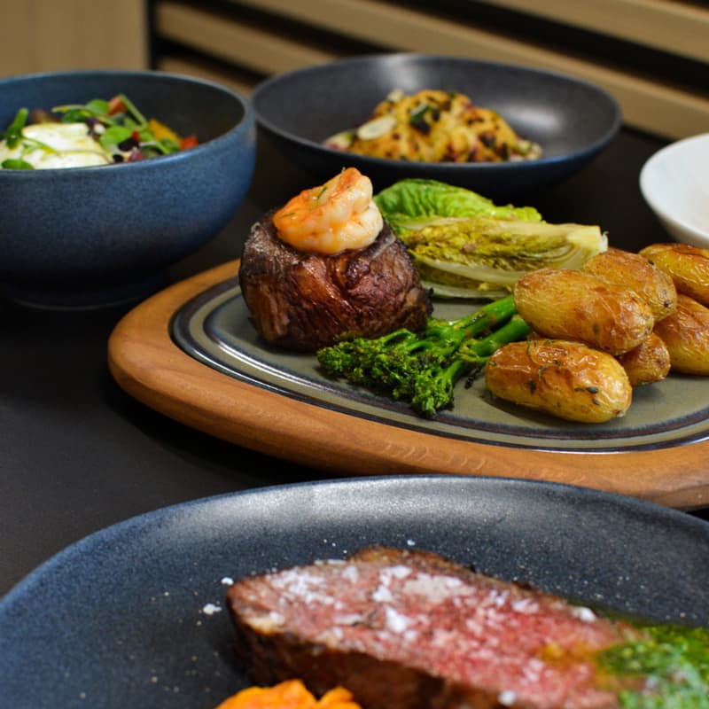 Steak s krevetami, pečenými bramborami, grilovaným salátem a brokolicí, doplněný salátem a těstovinami v misce.