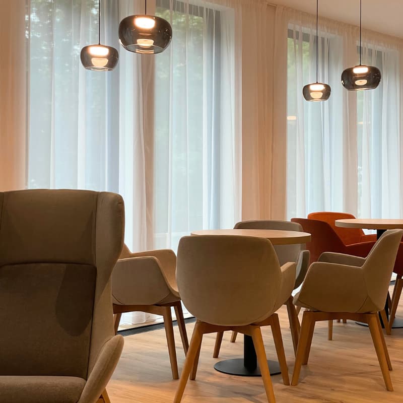 Moderní jídelna s béžovými a oranžovými čalouněnými židlemi kolem kulatých dřevěných stolů, závěsnými svítidly a velkými okny s průsvitnými závěsy.