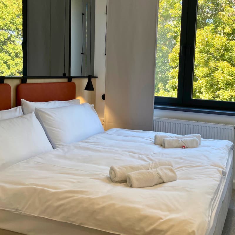 Úhledná manželská postel s bílými polštáři a složenými ručníky, umístěná vedle okna s výhledem na zelené stromy.