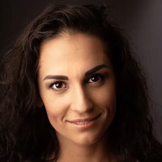 Detailní portrét usmívající se ženy s tmavými kudrnatými vlasy a neutrálním make-upem na tmavém pozadí.