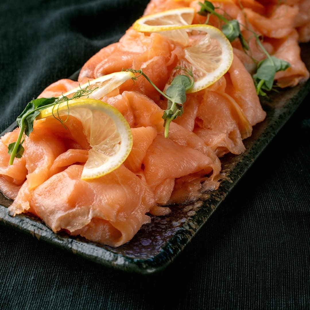 Talíř s tenkými plátky uzeného lososa, zdobený plátky citronu a čerstvými bylinkami na tmavém podkladu.