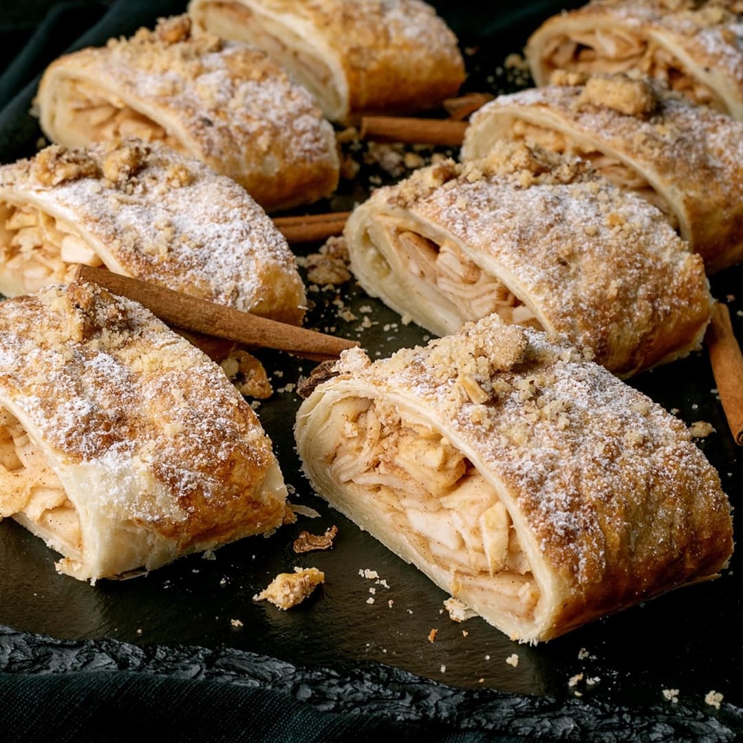 Plátky jablečného štrúdlu posypané moučkovým cukrem a streuselem na tmavém povrchu s tyčinkami skořice.
