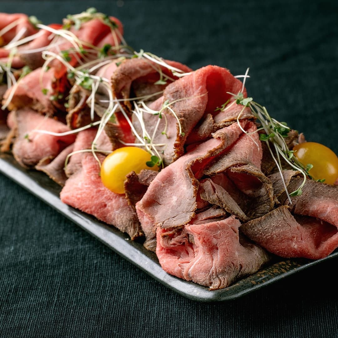 Tenké plátky krvavého roastbeefu zdobené klíčky a žlutými cherry rajčátky na obdélníkovém kovovém podnosu.