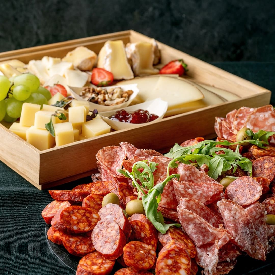 Dřevěný podnos s výběrem sýrů, hroznovým vínem, jahodami, vlašskými ořechy a marmeládou, vedle talíře s nakrájenými uzeninami, olivami a rukolou.