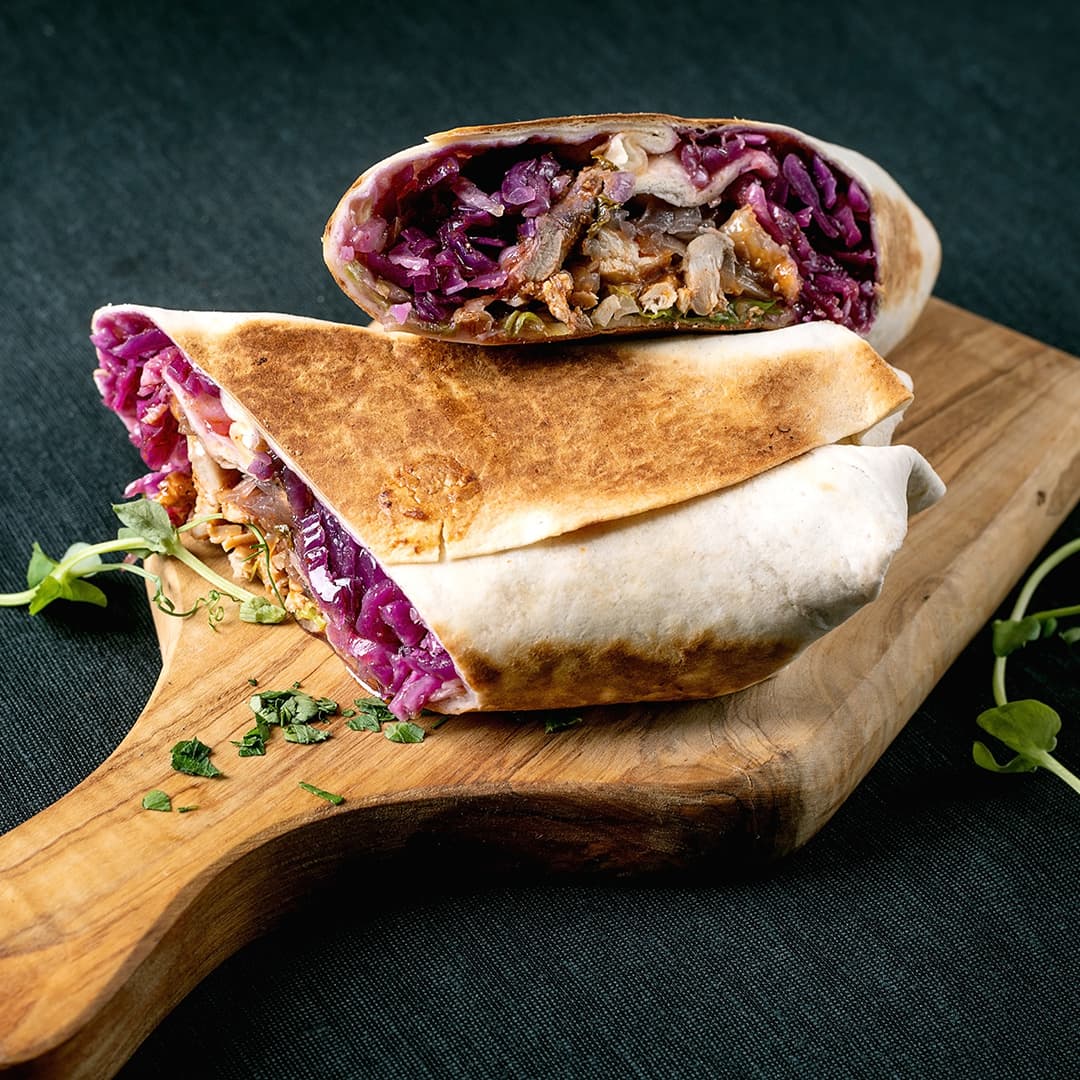 Grilovaný wrap rozkrojený na polovinu na dřevěné desce, plněný fialovým zelím, masem a zeleninou.