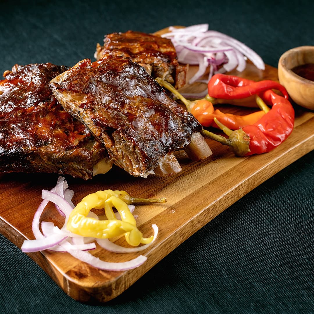Grilovaná žebra glazovaná omáčkou podávaná na dřevěném prkénku s nakrájenou červenou cibulí, žlutými a červenými chilli papričkami a barbecue omáčkou jako přílohou.