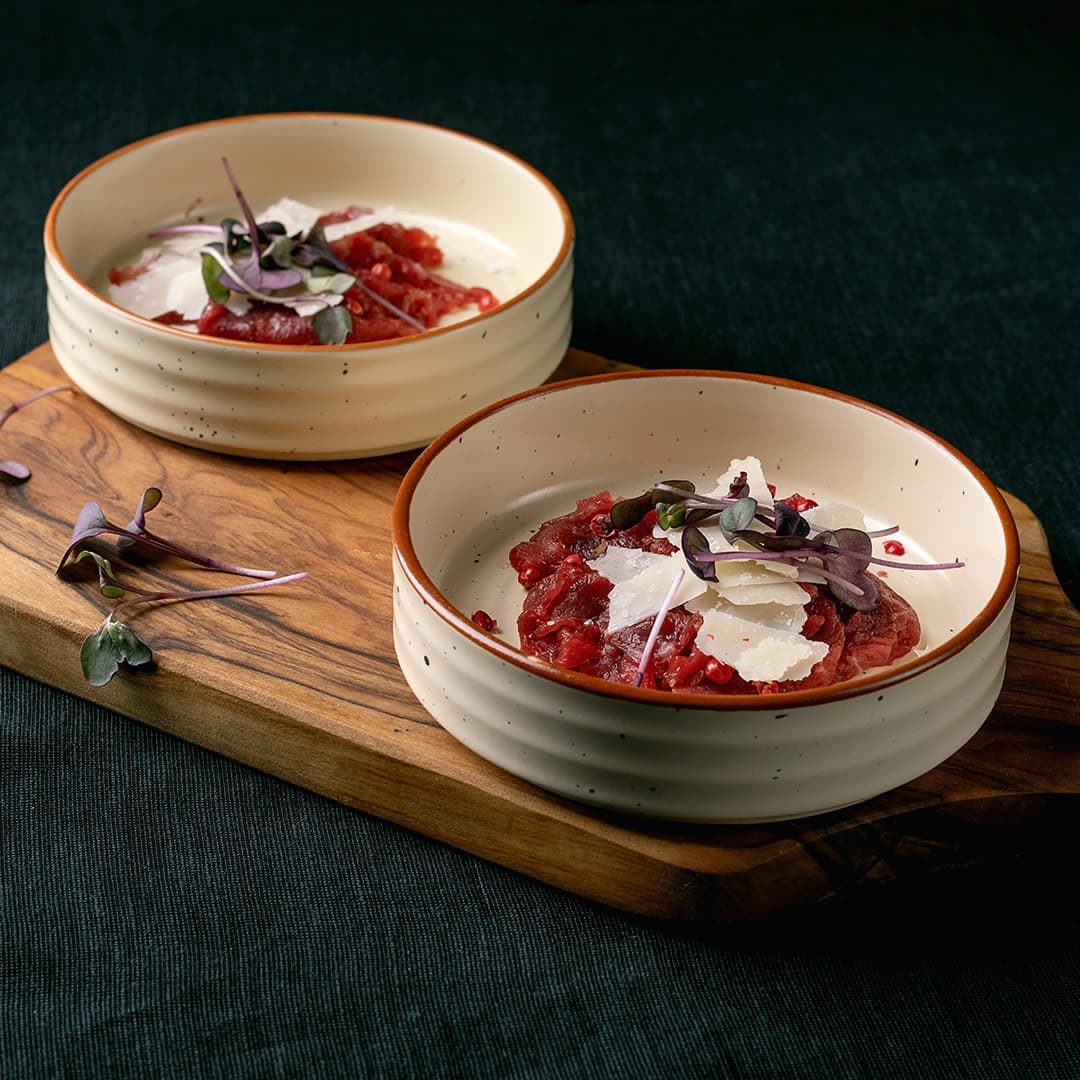 Dvě béžové misky s červeným tatarákem, posypané hoblinkami sýra a mikrozeleninou, podávané na dřevěném prkénku.