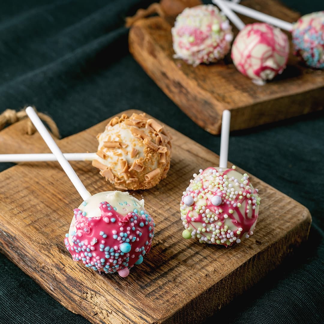 Tři barevné dortíky na špejli s posypem a čokoládovými kudrlinkami na dřevěné desce.