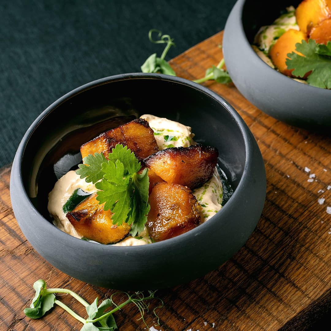 Černá mísa s kousky pečeného banánu na krémové omáčce, zdobená koriandrem, umístěná na dřevěném podkladu s rozsypanými bylinkami a vločkami soli.