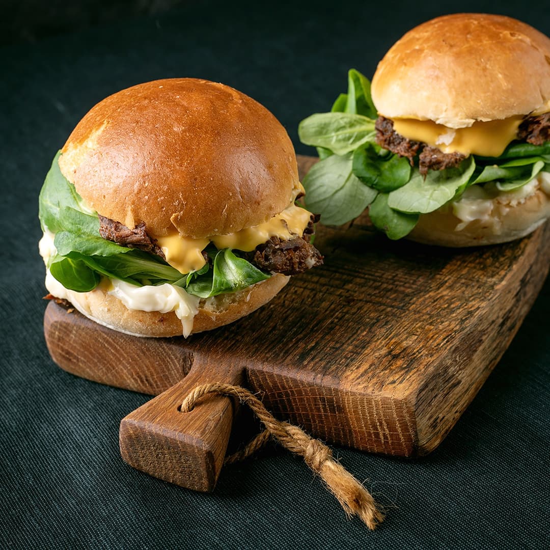 Dva cheeseburgery s listovou zeleninou na dřevěném podnosu na tmavém látkovém pozadí.