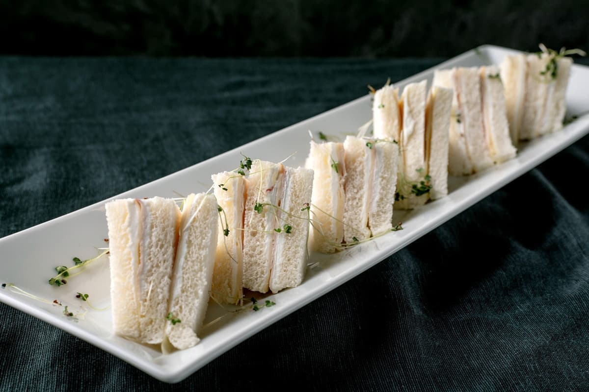 Obdélníkový bílý talíř s úhledně naservírovanými sendviči zdobenými malými zelenými klíčky na tmavém látkovém povrchu.
