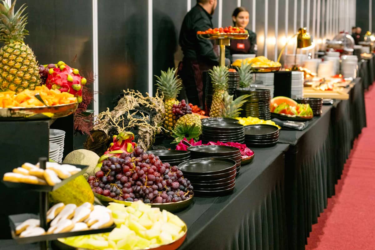 Dlouhý bufetový stůl s výběrem čerstvého ovoce, včetně ananasů, hroznů, dračího ovoce a melounů, spolu s naskládanými černými talíři a dvěma zaměstnanci ABF Catering v pozadí.