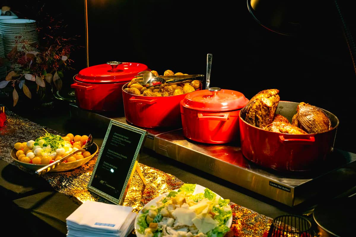 Bufetové občerstvení s červenými servírovacími miskami obsahujícími pečené maso a brambory, miskou cherry rajčat a kuliček mozzarelly a talířem Caesar salátu na černém stole se zlatým dekorem.