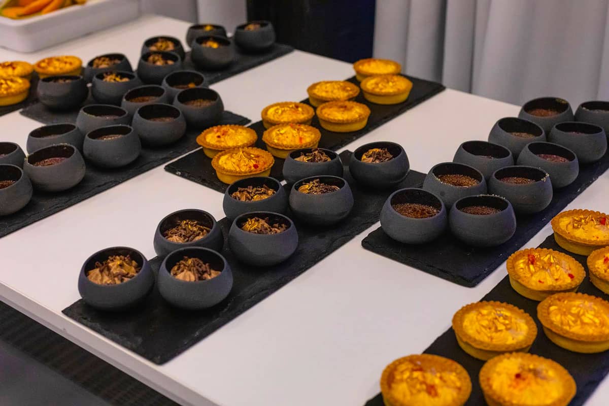 Řady malých černých kulatých dezertních košíčků a zlatých mini tartaletek na černých obdélníkových podnosech na bílém stole.